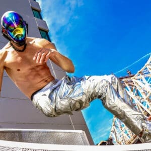 A masked man performs a high kick outdoors in shiny pants.